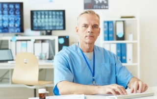 Urolog w niebieskim uniformie pracuje przy komputerze w gabinecie lekarskim.